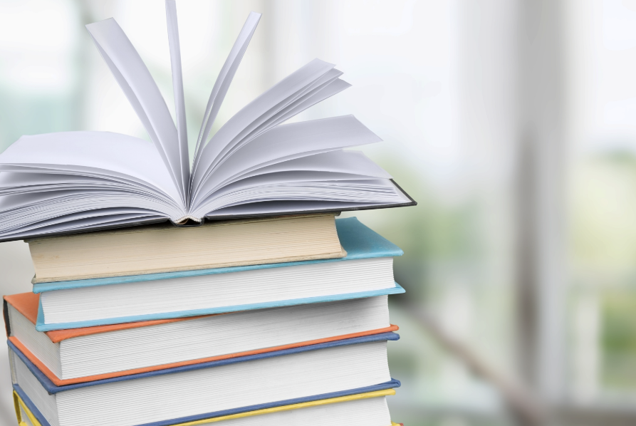 Ein aufgeschlagenes Buch mit aufgefächerten Seiten liegt auf einem Stapel verschiedenfarbiger Bücher vor einem hellen Hintergrund.
